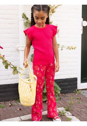 Jong meisje dat een felroze top en een broek met aardbeienprint draagt, houdt een geel gevlochten mandje vast buiten bij een witte muur.