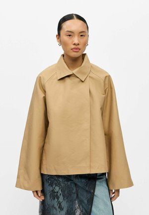 Femme aux cheveux foncés portant une veste beige à manches larges et un jean bleu avec un empiècement en dentelle florale noire sur une jambe, debout devant un fond uni.