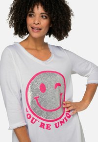 Weißes Langarmshirt mit einem großen silbernen Glitzer-Smiley-Gesicht mit rosa Akzenten und dem Text "DU BIST EINZIGARTIG" darunter. Texturierter Saum.