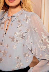 Ljust blå blus med en volangkrage, broderi av silverstjärnor och månar, genomskinliga ärmar och en knytning vid halsringningen.