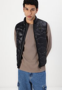 Svart polstret vest med kviltet design, frontglidelås og subtil logo-detalj. Bært over en beige langermet skjorte og blå denimbukser.