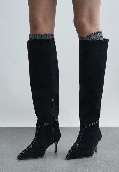 Μαύρες σουέντ μπότες μέχρι το γόνατο με μυτερή μύτη, ασημί ραφές και χρυσό φερμουάρ. Γκρι ραφιναρισμένες κάλτσες προεξέχουν από την κορυφή.