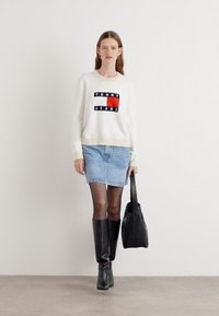 Hvit genser med marineblå og rød "TOMMY JEANS"-logo, blå denimskjørt, svarte kneehøye støvler og en svart totebag mot en lys bakgrunn.