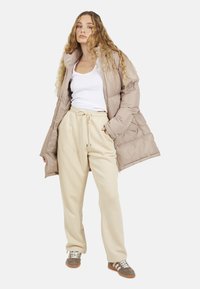 Beżowa kurtka puchowa z pikowanym wzorem, biały top na ramiączkach, kremowe dresy i szaro-brązowe sneakersy z białymi paskami. Gładka faktura na całej powierzchni.