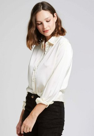 Blouse blanche en soie avec un devant à boutons, manches longues retroussées aux poignets, et un motif texturé subtil. Associée à un pantalon noir taille haute.
