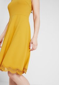 Žluté bezramínkové šaty s A-liniovým střihem, s krajkovým lemováním na spodním okraji a hladkým, texturovaným materiálem.