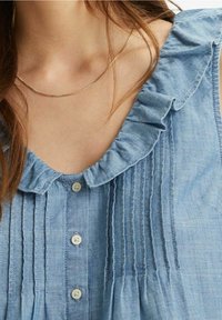 Närbild av en person som bär en ljusblå blus med volanger och en tunn guldhalsband runt halsen.