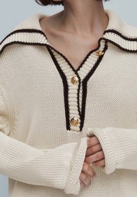 Personne portant un pull en maille crème avec bordure noire et boutons dorés, les mains partiellement couvertes par des manches longues, tenues ensemble à la taille.
