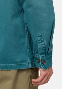 Giacca in denim verde acqua con polsini a bottoni, cuciture visibili e taglio dritto. Texture liscia con una vestibilità strutturata.