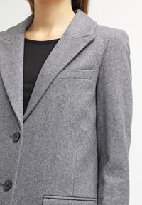 Blazer grigio in lana con revers a punta, due bottoni frontali e una tasca sul petto; la texture appare liscia con una finitura morbida.