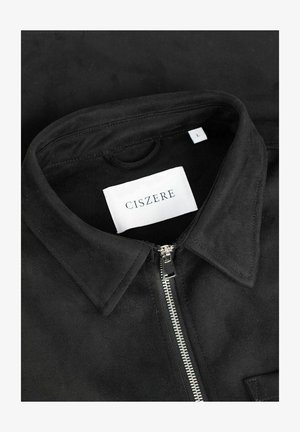 Svart semsket skinnjakke med glidelås foran, spiss krage og etikett som sier "CISZERE." Har en sidelomme og glatt tekstur.