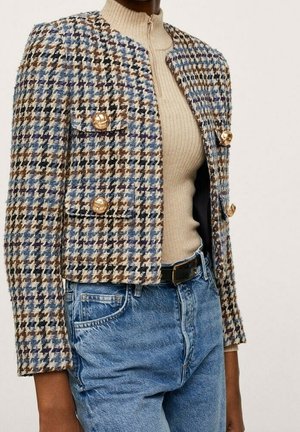 Kobieta w krótkiej kurtce w pepitkę z złotymi guzikami, nosząca beżowy prążkowany golf i niebieskie dżinsy z wysokim stanem oraz czarnym paskiem.