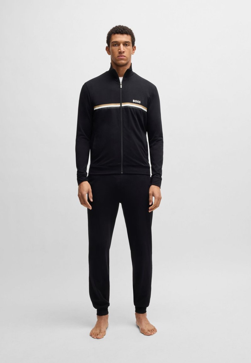 Schwarze Zip-Up-Trainingsjacke mit weißen und goldenen horizontalen Streifen und Logo, kombiniert mit passenden schwarzen Jogginghosen, beide aus glattem Stoff.