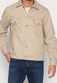 Veste en denim beige avec col, deux poches poitrine et fermeture par boutons. Caractérisée par des éléments métalliques argentés et des détails de coutures.