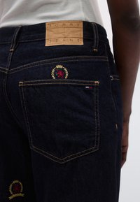 Tumed sinist värvi teksapüksid, millel on nahast silt „TOMMY JEANS“ ning tikitud lõvi ja tammelehe detailid tagataskul.