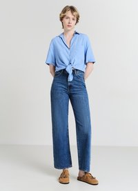 Chemise à manches courtes bleu clair nouée à la taille, associée à un jean en denim bleu foncé à jambes droites. Les accessoires comprennent des mocassins en daim beige.
