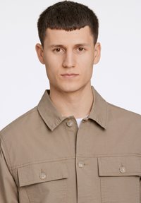 Beige button-up jacket featuring a spread collar, two chest pockets, and a smooth texture. Minimalist design with a relaxed fit.
