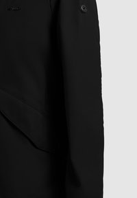 Must jopeserv, millel on nööbi detail ja osaliselt nähtav klapiga tasku, ühel ühtlasel heledal taustal.