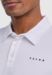 Biała koszulka polo wykonana z teksturowanej tkaniny, z ostrym kołnierzykiem, trzema guzikami oraz czarnym logo "FALKE" wyszytym na piersi.