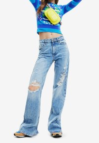 Lichtblauwe versleten denim jeans met brede pijpen, voorzien van grote scheuren op beide knieën. Gecombineerd met een aansluitend topper in blauwe en groene strepen en een gele tas.