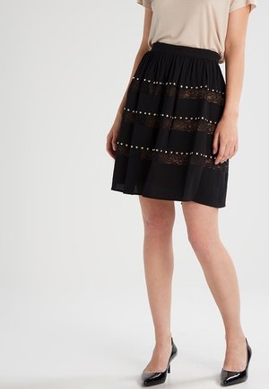 Femme portant une jupe noire longueur genou avec de la dentelle et des rayures horizontales cloutées, haut beige et chaussures à talons noirs, debout.