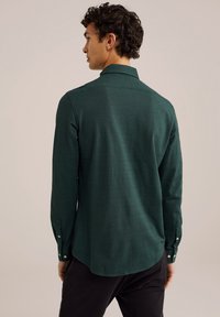 Majica s dolgimi rokavi v temno zeleni tkanini, ki ima klasičen ovratnik, gumbnice na rokavih in rahlo ukrivljen rob. Gladka tekstura z rahlim tkanjem.