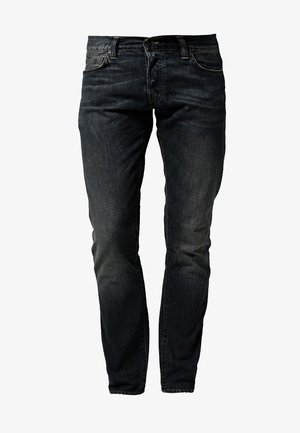 Homme portant un débardeur rouge, un jean foncé, des baskets blanches, tenant des lunettes de soleil et portant un sac à dos sur une épaule.