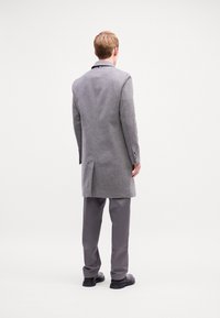 Cappotto di lana grigio con un design sartoriale, aperture posteriori singole e dettagli in bottone. Abbinato a pantaloni grigi e scarpe nere.