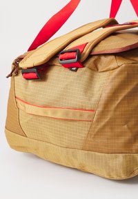 Duffle bag in yellow mesh fabric with red accents, featuring a flap closure, dual side clips, and a front zip pocket. Textured surface detail.
