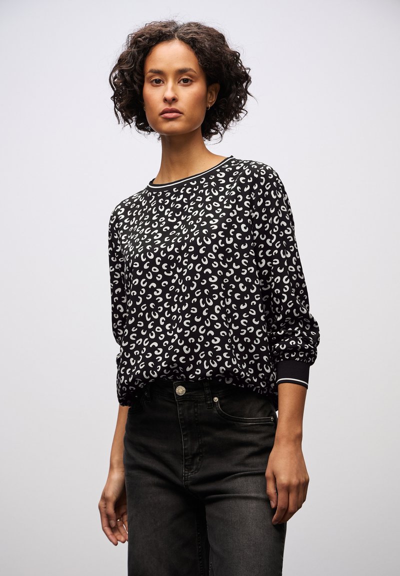Donna con capelli corti e ricci che indossa una maglietta a maniche lunghe con un motivo in bianco e nero e jeans a vita alta scuri, in piedi contro uno sfondo neutro.