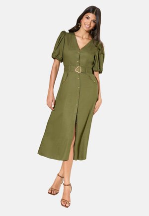 Žena oblečená v olivovo zelených midi šatách na gombíky s naberanými rukávmi a opaskom, doplnená hnedými sandálmi na vysokom opätku so remienkami.
