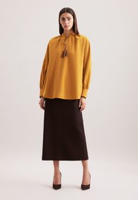 Senfgelbe Bluse mit hohem Kragen und Kordelzug, kombiniert mit einem dunkelbraunen Midikleid und dunklen High Heels.