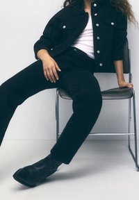 Zwarte spijkerjasje met zilverkleurige knopen, gecombineerd met een zwarte broek. Model draagt een wit topje en zwarte laarzen, zittend op een metalen stoel.