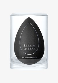 Spugna per il trucco in schiuma nera all'interno di un contenitore cilindrico trasparente. Presenta una forma a uovo con una superficie liscia ed è etichettata con le istruzioni per l'uso.