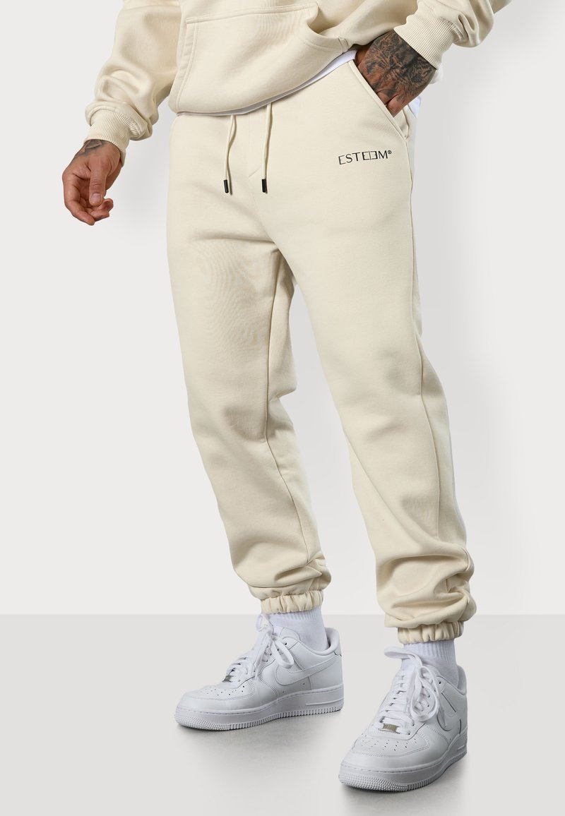 Crèmefarbene Jogginghosen aus weichem Stoff mit elastischen Bündchen, auf der Vorderseite mit einem kleinen "ESTEEM"-Logo. Kombiniert mit weißen Sneakers und Crew-Socken.