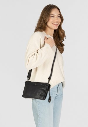 Bolso de cuero negro tipo bandolera con cierre de cremallera, textura suave y correa ajustable, llevado por una persona con un suéter crema y jeans claros.