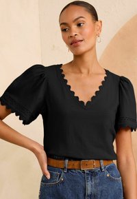 Femme portant un haut noir à col en V festonné avec des manches bouffantes et un jean bleu avec une ceinture brune, se tenant devant un mur beige.