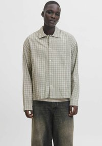 Leichte, karierte Button-Down-Bluse mit grünen, braunen und weißen Farbtönen, entspannter Schnitt und einer Brusttasche. Über dunkler Jeans getragen.