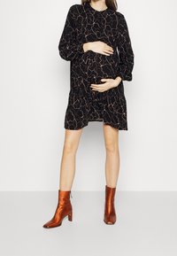 Schwarzes Kleid mit abstraktem, beigem Muster, lockerer Schnitt und langen Ärmeln; kombiniert mit glänzenden, kurz geschnittenen, orangen Stiefeletten.