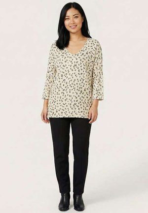 Femme debout, portant un haut beige à manches longues imprimé léopard, un pantalon noir et des bottines noires, souriante sur un fond uni.