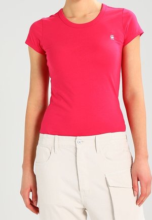 Femme portant un t-shirt ajusté rose vif à manches courtes et un pantalon beige clair avec poches avant et fermeture par bouton.