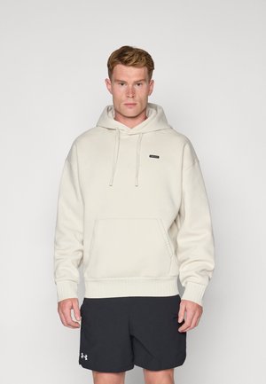 Cream hoodie with a front pocket, drawstring hood, ribbed cuffs, and hem. Worn with black shorts, features a small brand label on the chest.
