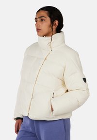 Giacca puffer crema con colletto alto, chiusura con zip e tasche laterali. Presenta un emblema nero sulla manica e un motivo trapuntato per texture.