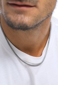 Collana in metallo con design intrecciato, finitura color argento, posata su una maglietta bianca, che esalta il collo di chi la indossa.