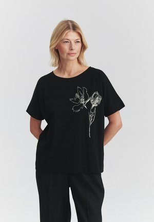Donna bionda che indossa una maglietta nera a maniche corte con un disegno floreale bianco sul davanti, in piedi davanti a uno sfondo chiaro uniforme.
