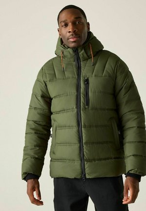 Mężczyzna ubrany w oliwkowo zieloną pikowaną kurtkę z kapturem i czarne spodnie, stojący na jasnym, jednolitym tle.