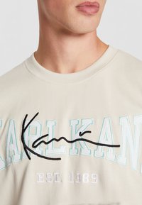 Sweatshirt beige clair avec le logo brodé "KARL KANI" en turquoise et noir, accompagné de la date de création "EST. 1989" en blanc. Texture douce.