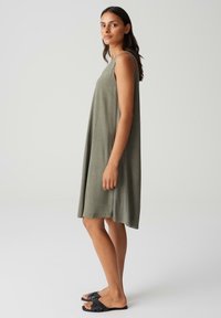 Sleeveless, knee-length dress in olive green fabric with a loose fit, featuring a rounded neckline and soft texture. Worn with black slide sandals.