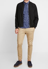 Giacca bomber nera sopra una camicia floreale blu, pantaloni slim-fit color khaki e scarpe in suede blu navy. Tessuto liscio con un design classico.