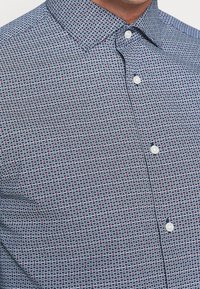 Nappisuljettava paita, jossa sinivalkoinen geometrinen kuvio, levitetty kaulus, valkoiset napit ja sileä kankaan rakenne.